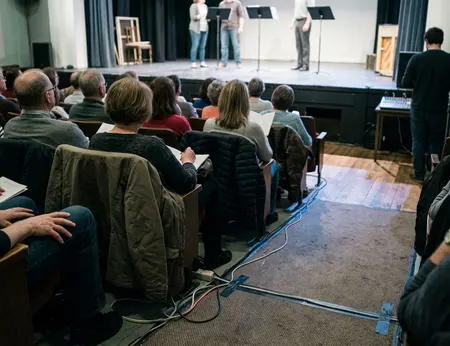 Audience scene at live event, showing rows of attendees watching a rehearsal-room musical run-through